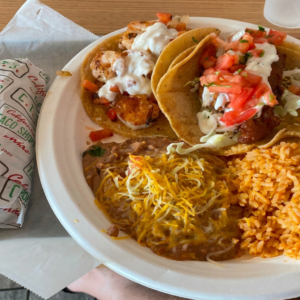 Plate of tacos topped with pico de gallo and cream, served with refried beans, melted cheese, and Mexican rice.